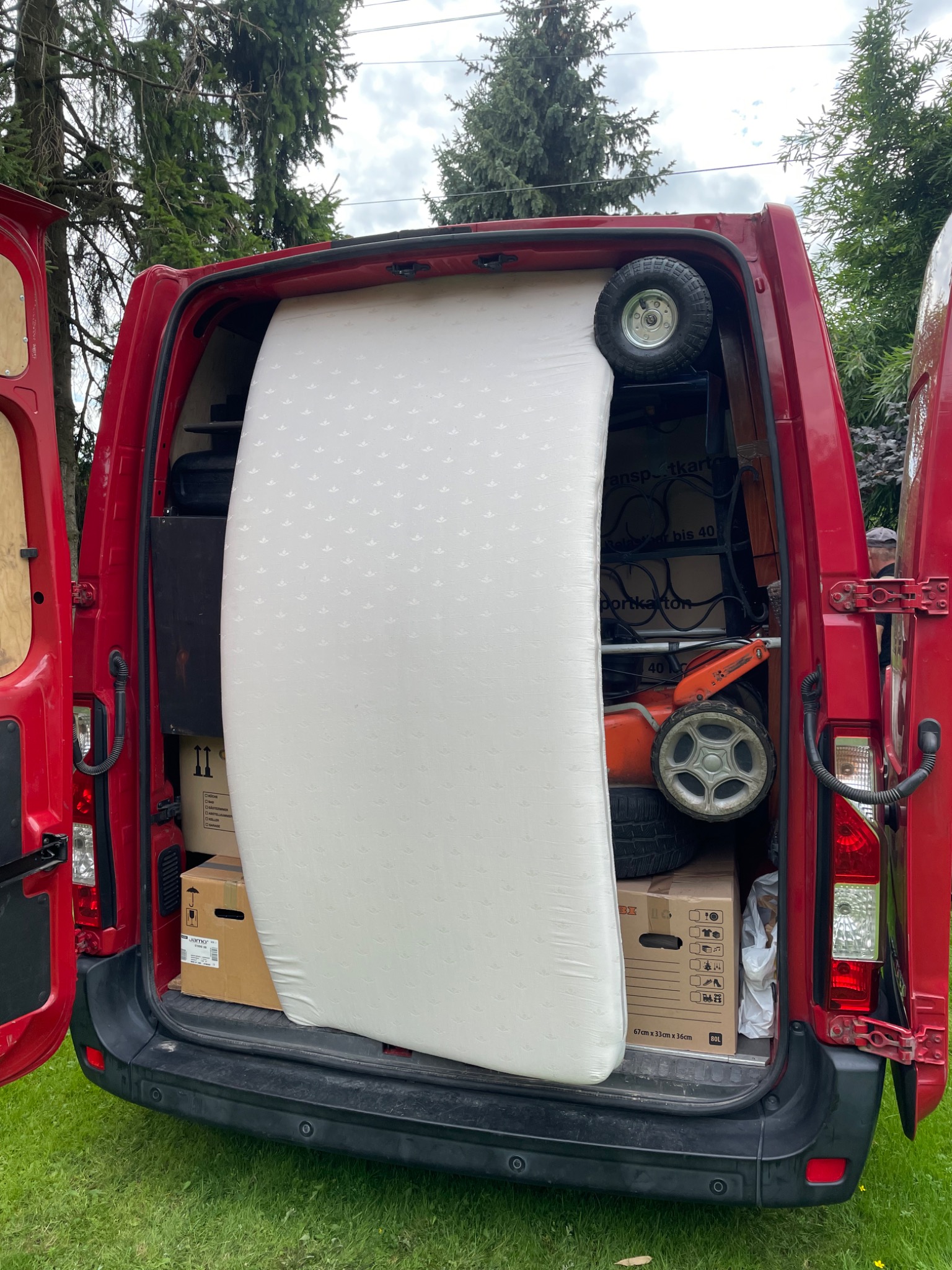 Wnętrze czerwonego busa z otwartymi drzwiami załadowane kartonami, materacem w pokrowcu i kosiarką do trawy podczas przeprowadzki.