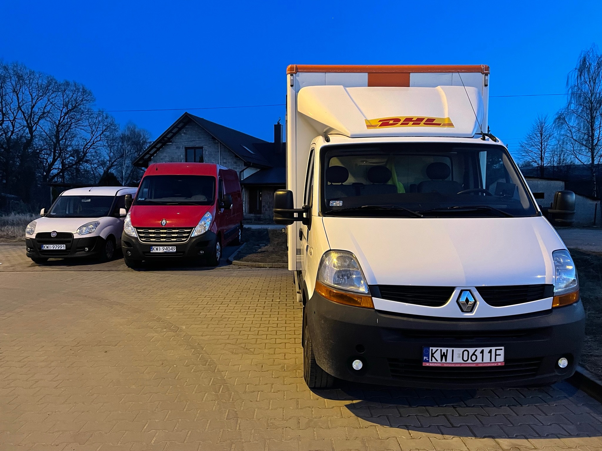 Trzy busy różnych marek (Fiat, Renault, DHL) zaparkowane na brukowanej powierzchni przed domem o zmierzchu.