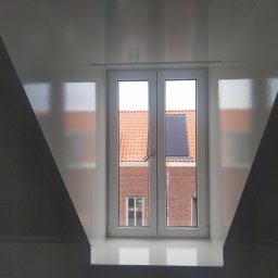 Artur Walenciuk - Białe, świeżo pomalowane poddasze z oknem, przez które widać ceglany budynek z pomarańczową dachówką i panelem solarnym.