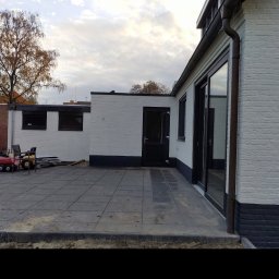 Artur Walenciuk - Świeżo pomalowana na biało elewacja budynku z ciemną podmurówką, widoczny taras z szarych płyt, drabina oparta o ścianę i zabawki dziecięce na placu budowy.