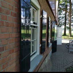 Artur Walenciuk - Ceglana elewacja budynku z oknami i czarnymi, błyszczącymi okiennicami, częściowo otwartymi, widok z boku, na chodniku leżą płyty