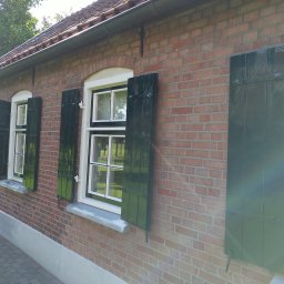 Artur Walenciuk - Ceglana elewacja budynku z oknami z białymi ramami i ciemnozielonymi okiennicami, częściowo otwartymi.