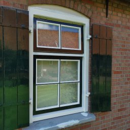 Artur Walenciuk - Okna z drewnianymi, zielonymi okiennicami na tle ceglanej ściany. Widoczne odbicie dachu w szybach.