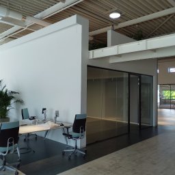 Artur Walenciuk - Nowoczesne biuro z białymi ścianami, szklanymi drzwiami, roślinami doniczkowymi i biurkami z krzesłami w kolorze turkusowym, przygotowywane do użytku, z widocznymi foliami ochronnymi na monitorach.