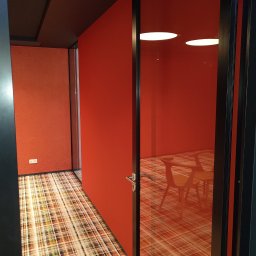 Artur Walenciuk - Pomarańczowy pokój biurowy z przeszklonymi ścianami i wzorzystą wykładziną, widok przez otwarte drzwi z czarną futryną.