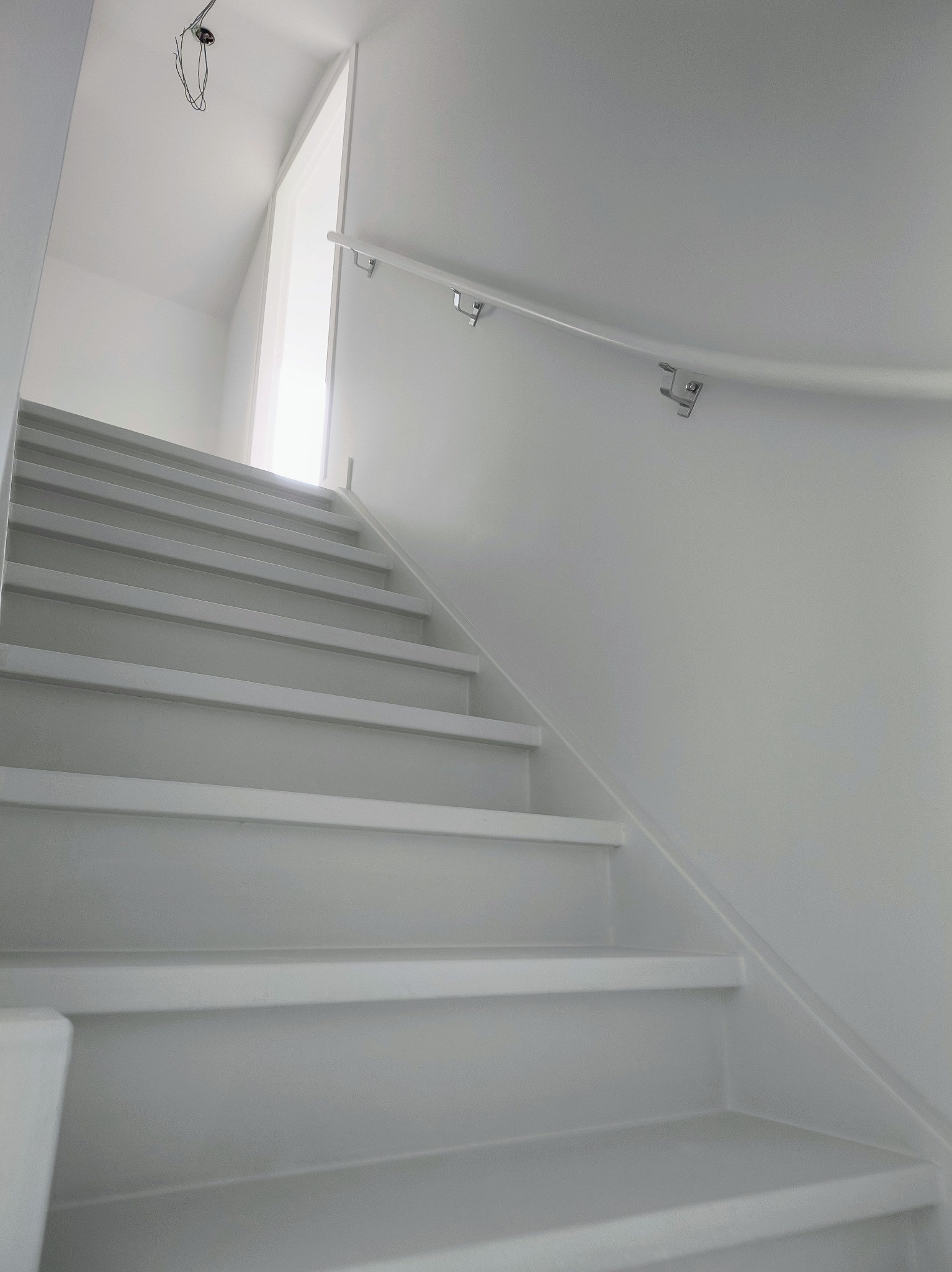 Białe schody z poręczą na białej ścianie, widoczne przewody elektryczne na suficie, minimalistyczne wnętrze.