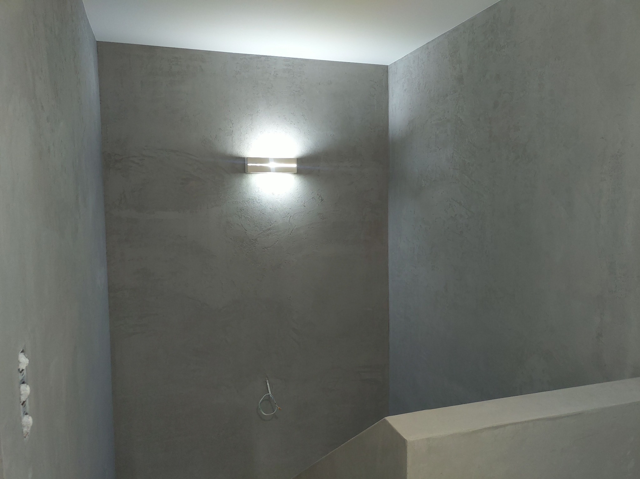 Szpachlowana ściana w odcieniach szarości z nowoczesnym podświetlanym kinkietem, widoczne niedokończone gniazdka elektryczne i fragment schodów.