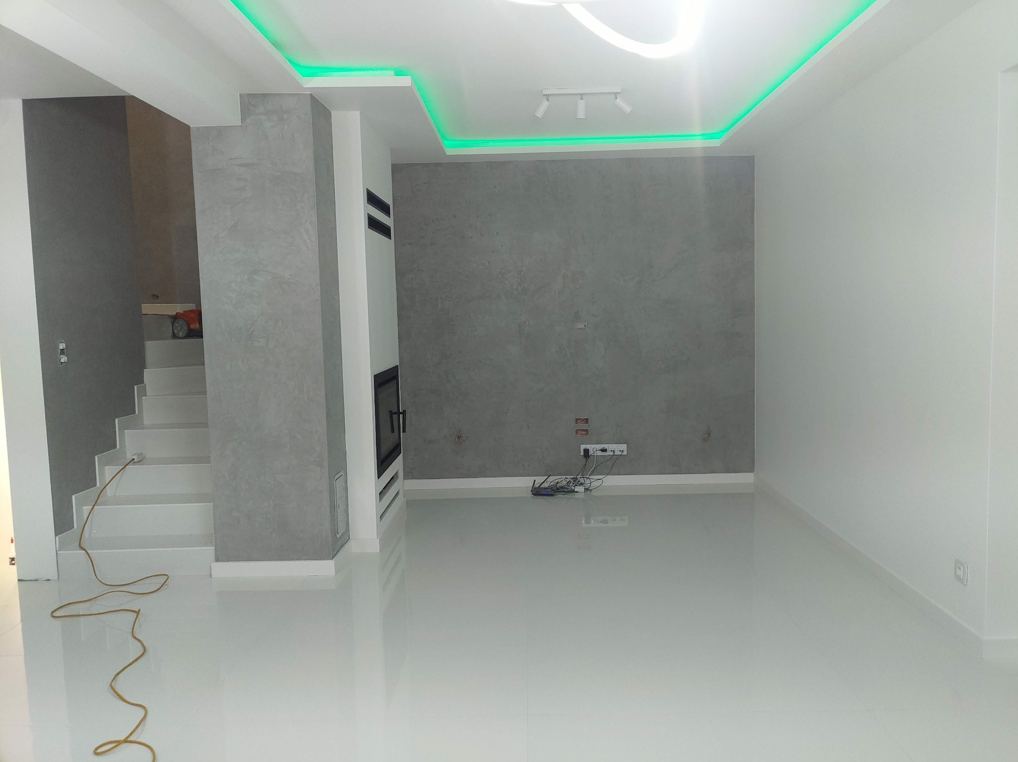 Wnętrze nowocześnie wykończonego salonu z szarą ścianą dekoracyjną, białymi podłogami i sufitem z zielonym oświetleniem LED, widoczne schody i kabel na podłodze.