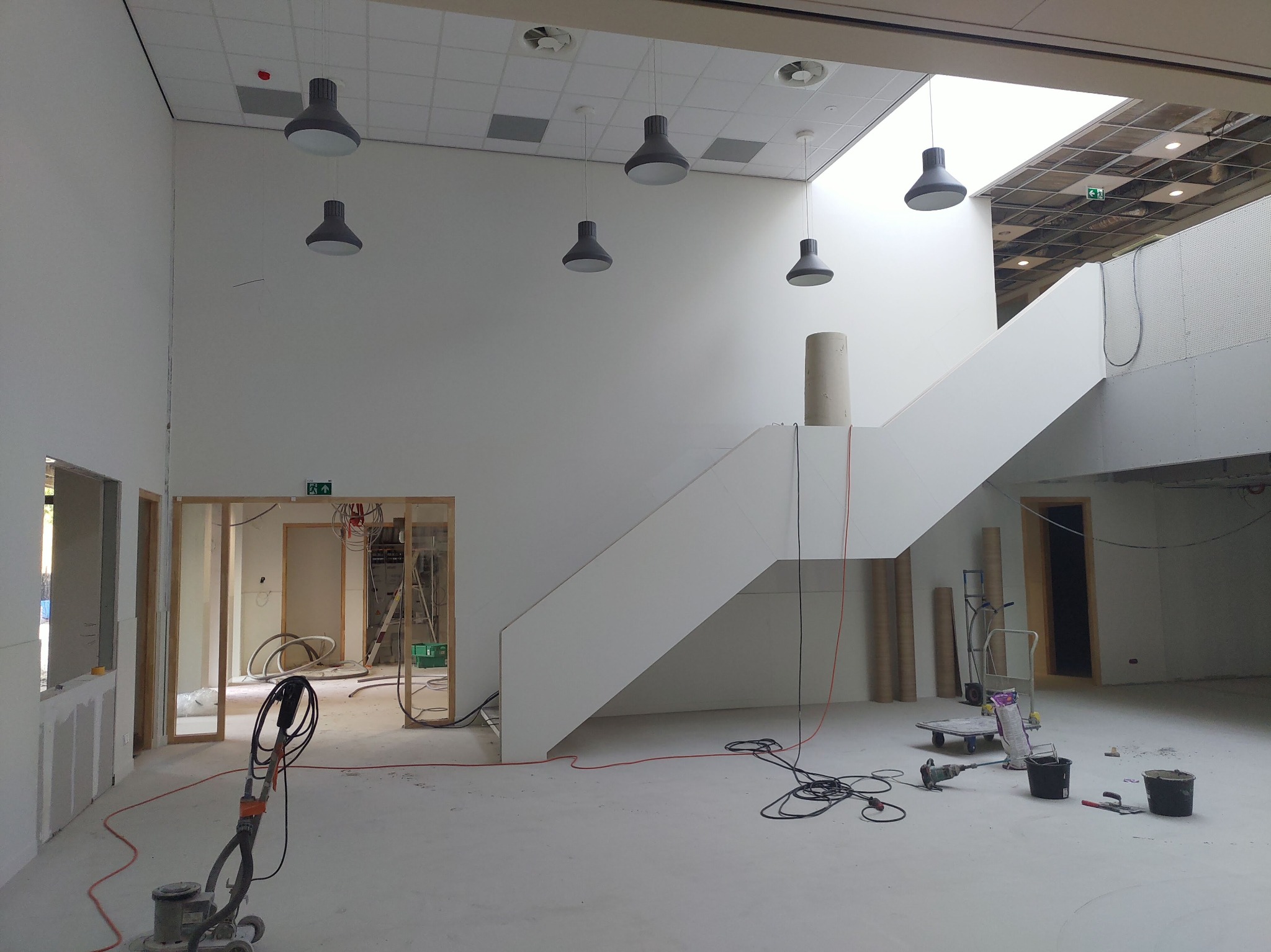 Wnętrze w trakcie prac wykończeniowych: białe ściany, schody, wiszące lampy, maszyna do polerowania podłóg, widoczne kable i narzędzia, surowy stan.