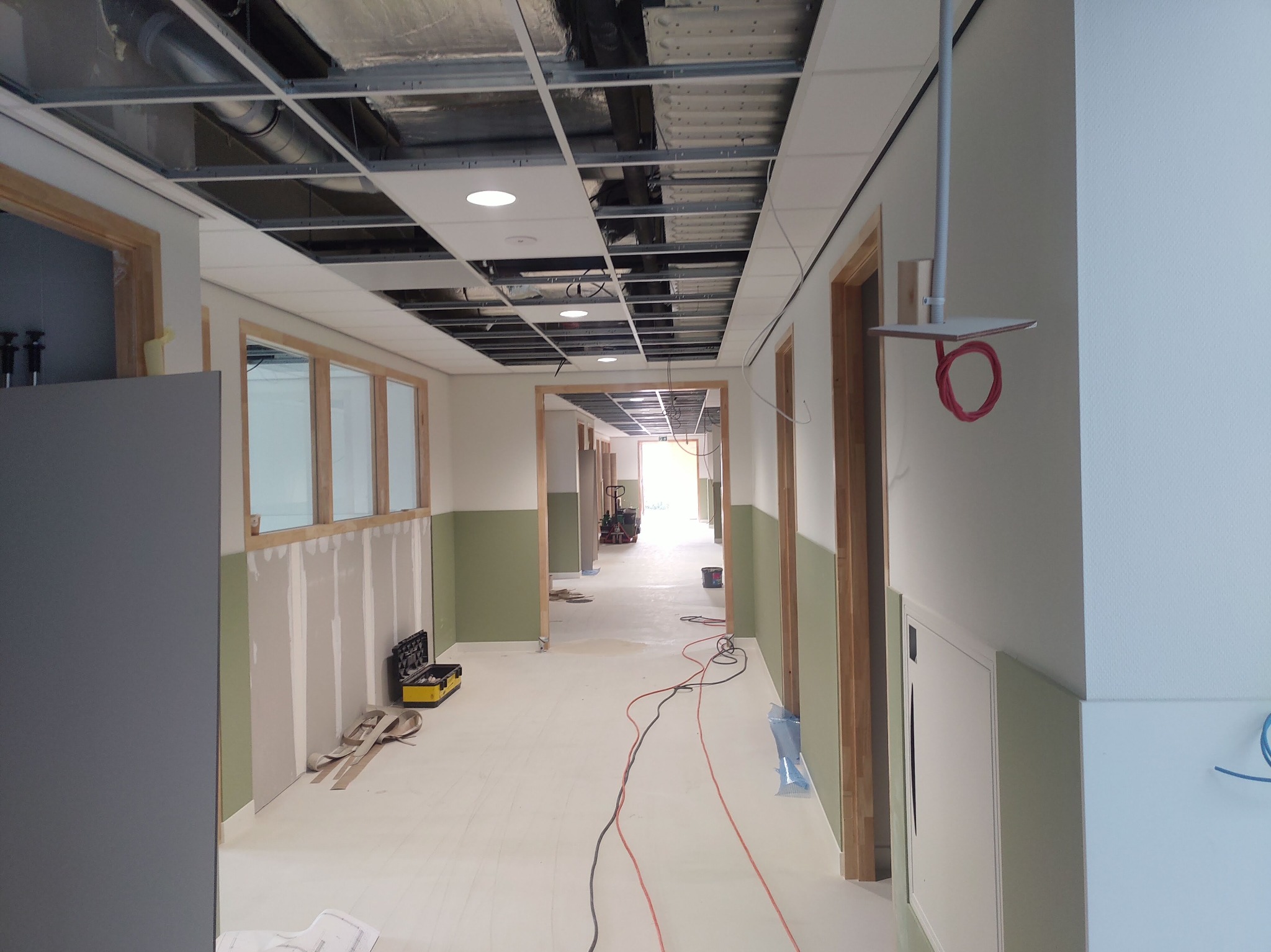 Korytarz w trakcie prac wykończeniowych: widoczne niezabezpieczone ściany z płyt gipsowo-kartonowych, częściowo pomalowane na zielono, sufit podwieszany w trakcie montażu, narzędzia i kable...
