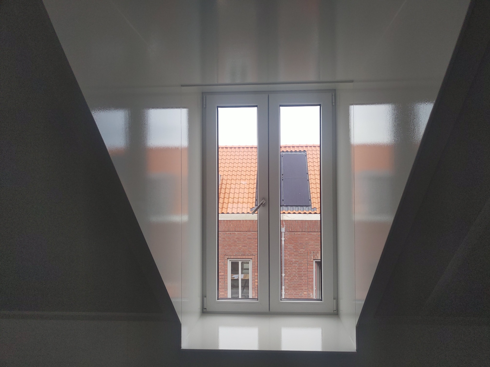 Białe, świeżo pomalowane poddasze z oknem, przez które widać ceglany budynek z pomarańczową dachówką i panelem solarnym.