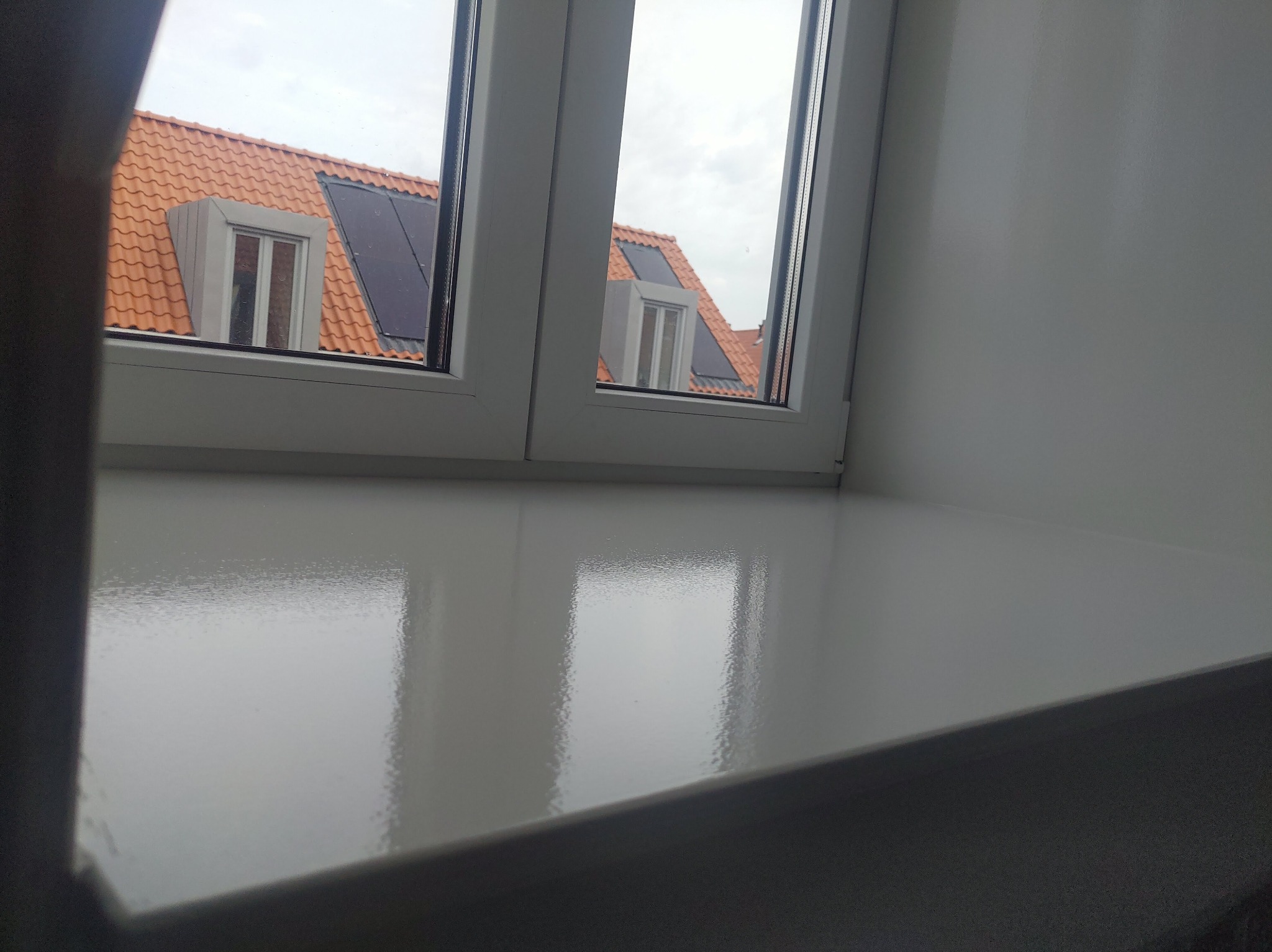 Błyszczący, świeżo pomalowany biały parapet wewnętrzny z widokiem na dach z czerwoną dachówką i panele słoneczne za oknem.