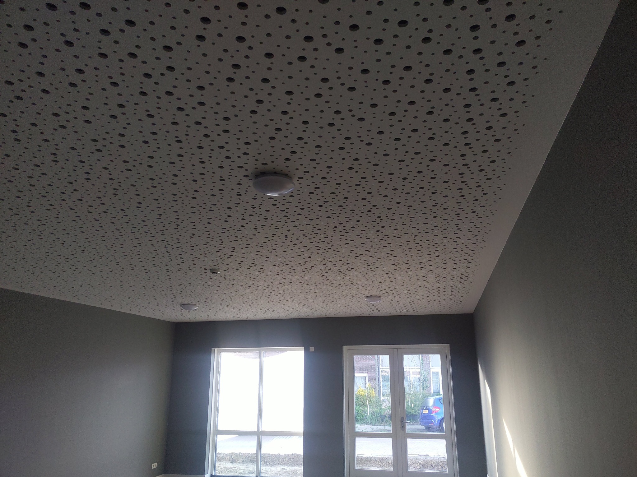 Szary sufit z perforacją w nowym pomieszczeniu, widoczne okna z zewnątrz i minimalistyczne oświetlenie.