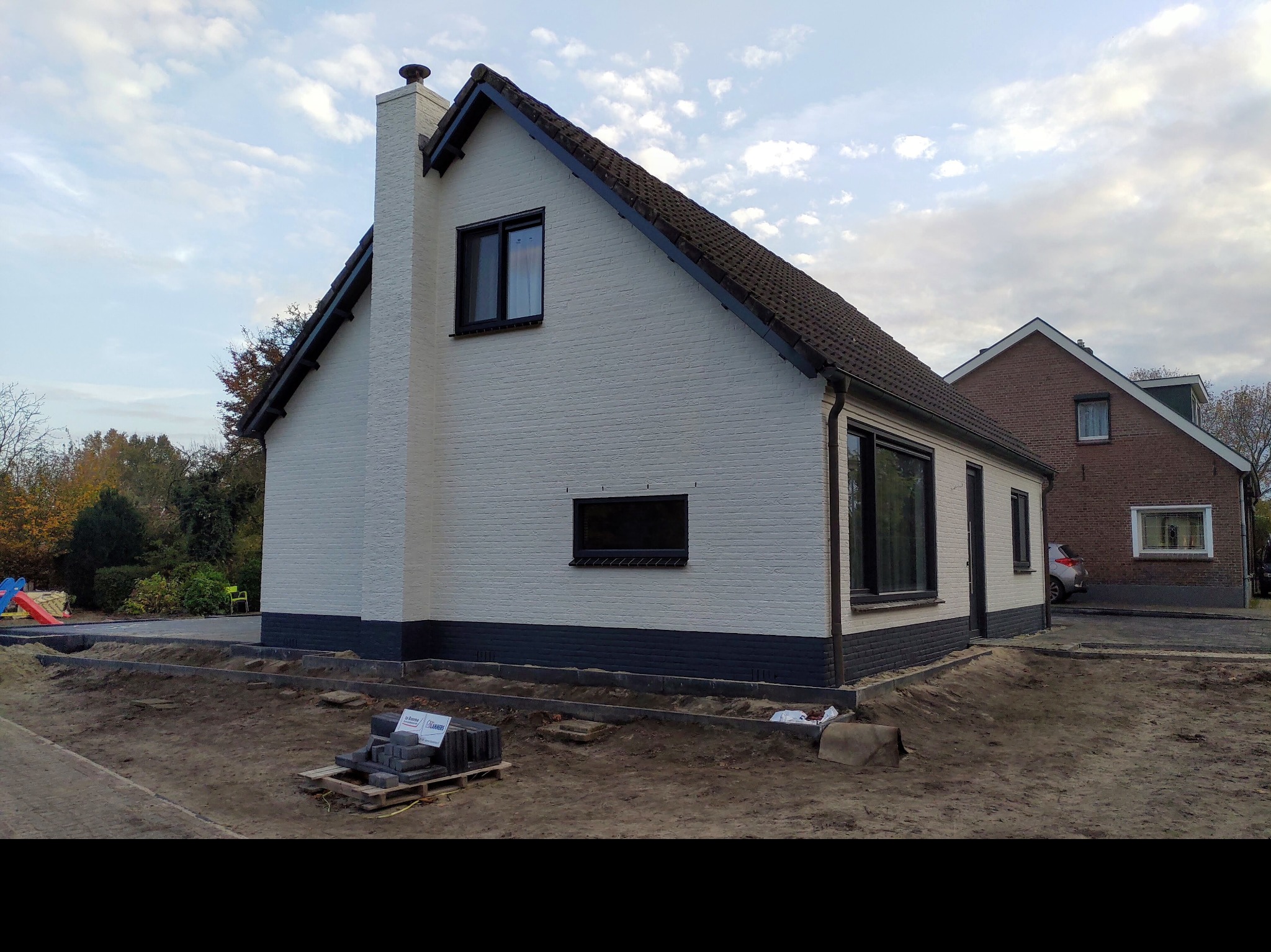Dwukondygnacyjny dom z białej cegły z czarnymi ramami okiennymi i ciemnoszarą podmurówką, widoczny komin, teren wokół domu przygotowany pod prace brukarskie, paleta z kostką brukową.