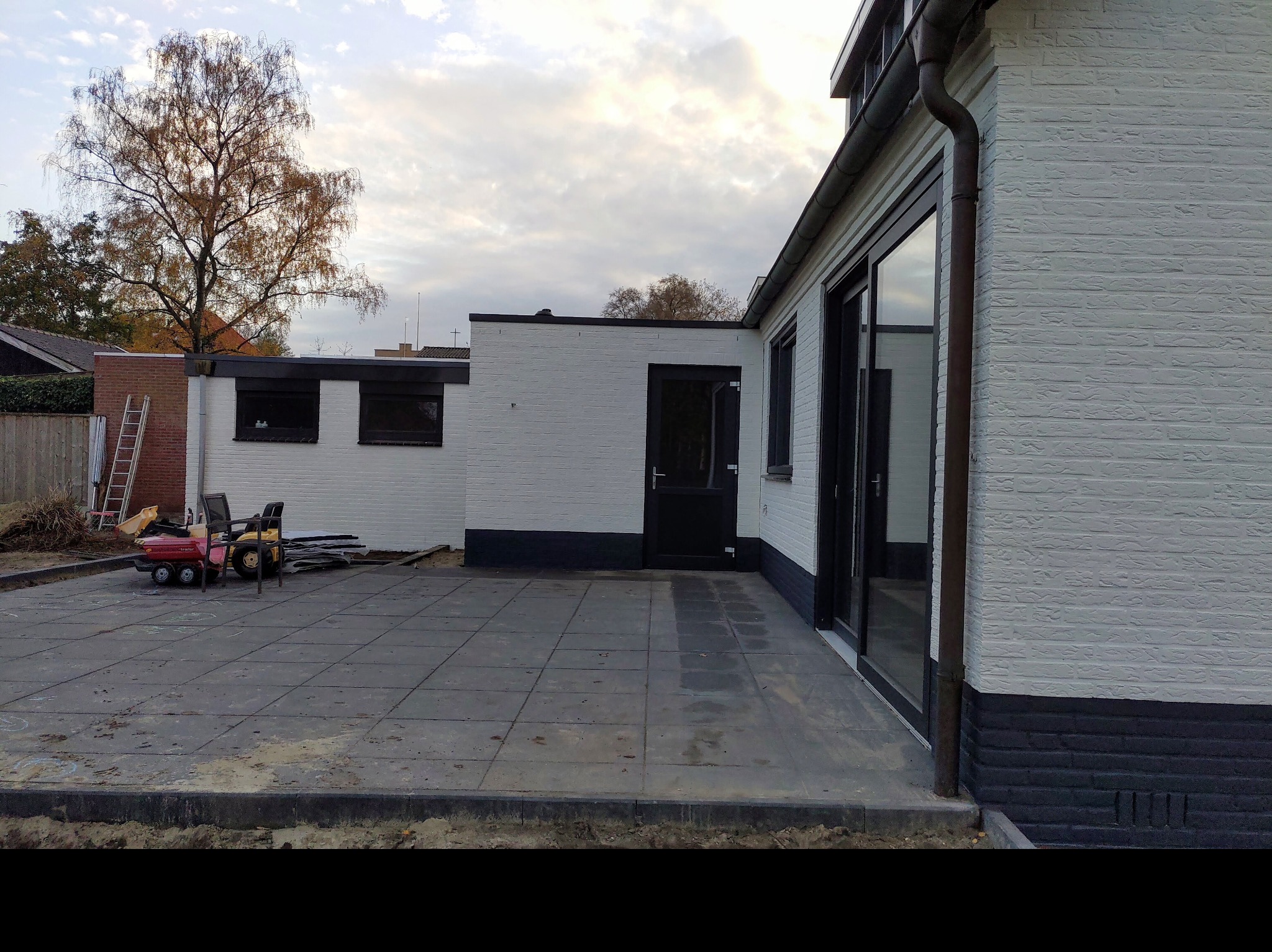 Świeżo pomalowana na biało elewacja budynku z ciemną podmurówką, widoczny taras z szarych płyt, drabina oparta o ścianę i zabawki dziecięce na placu budowy.