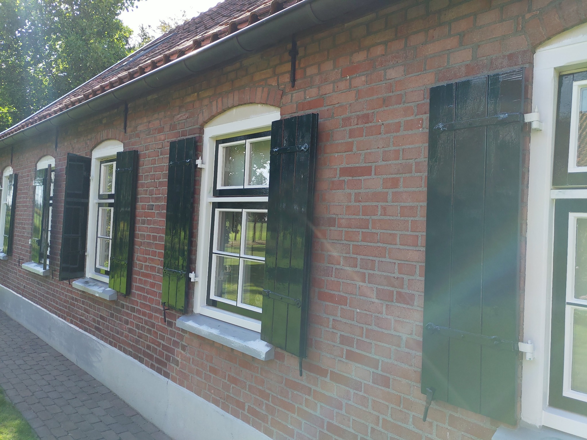 Ceglana elewacja budynku z oknami z białymi ramami i ciemnozielonymi okiennicami, częściowo otwartymi.