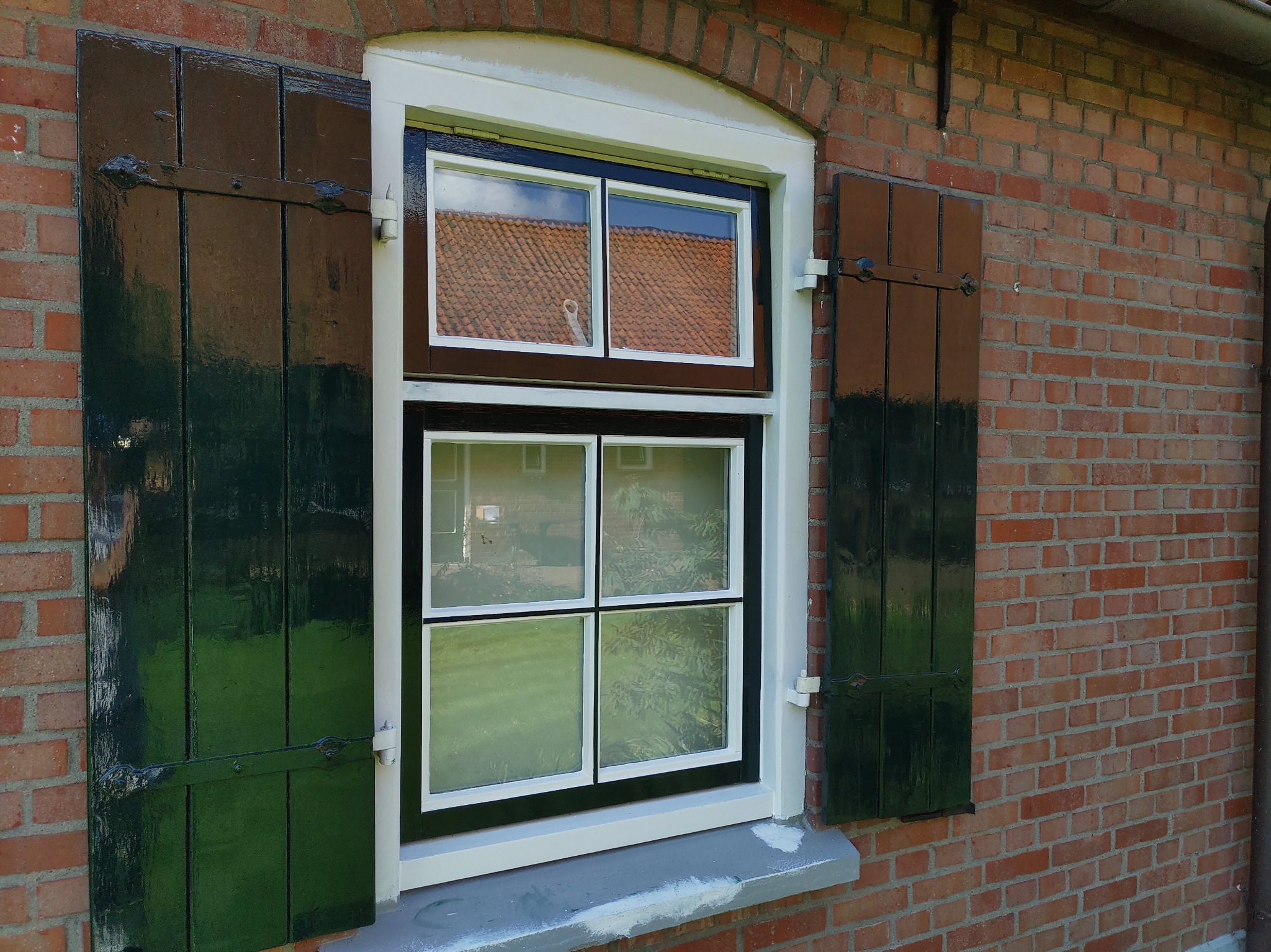 Okna z drewnianymi, zielonymi okiennicami na tle ceglanej ściany. Widoczne odbicie dachu w szybach.