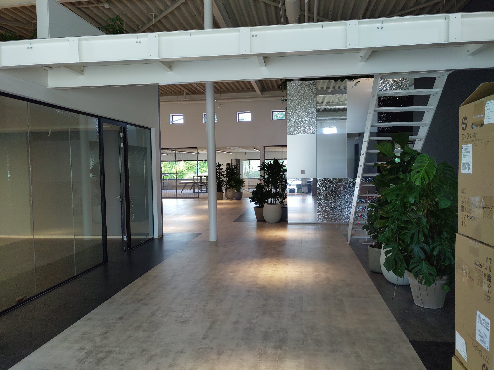 Wnętrze biura po remoncie: jasna podłoga imitująca kamień, szklane ścianki działowe, białe elementy konstrukcyjne sufitu i filar obłożony mozaiką szklaną, rośliny doniczkowe i kartony z monitorami.