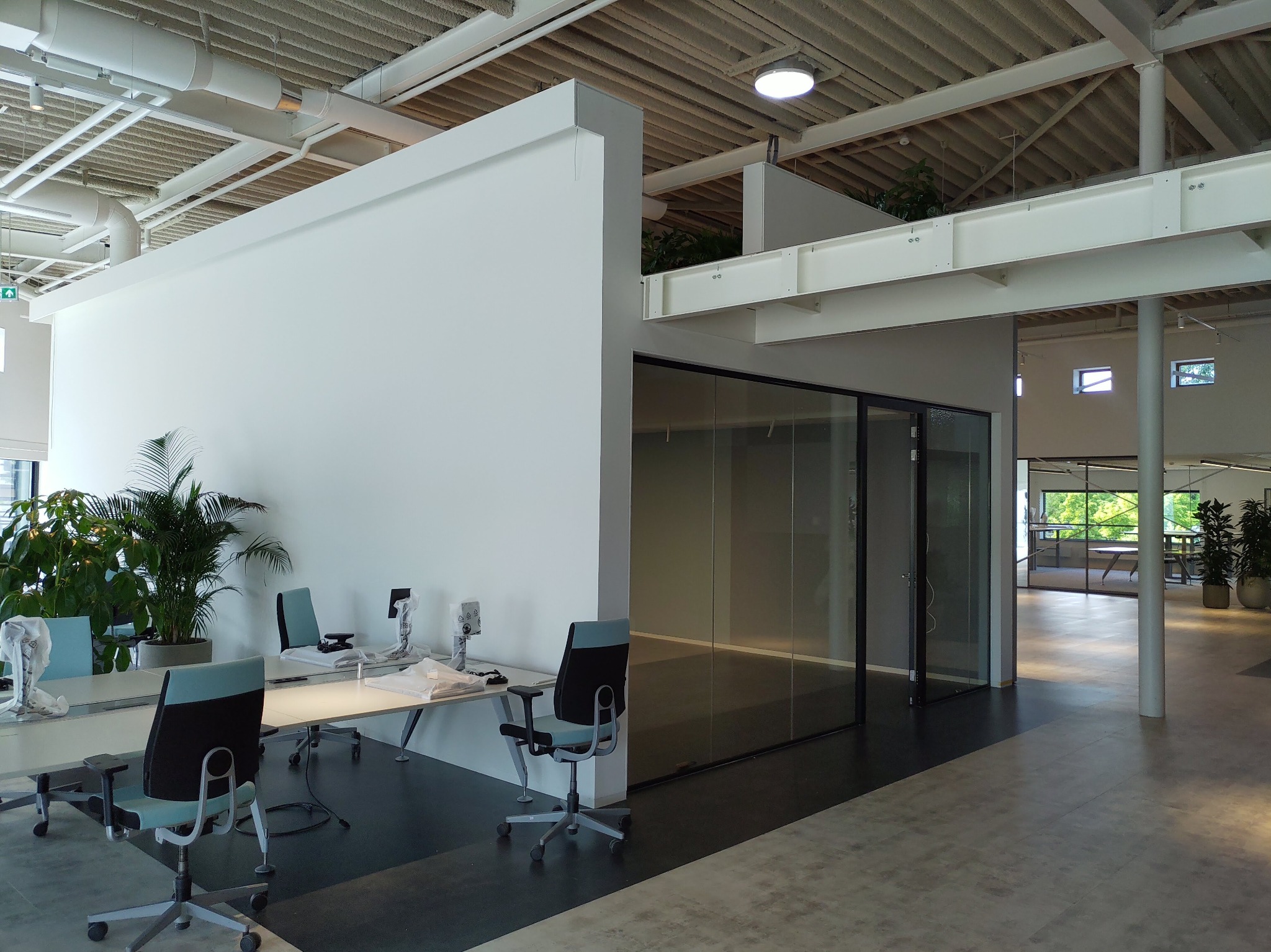 Nowoczesne biuro z białymi ścianami, szklanymi drzwiami, roślinami doniczkowymi i biurkami z krzesłami w kolorze turkusowym, przygotowywane do użytku, z widocznymi foliami ochronnymi na monitorach.