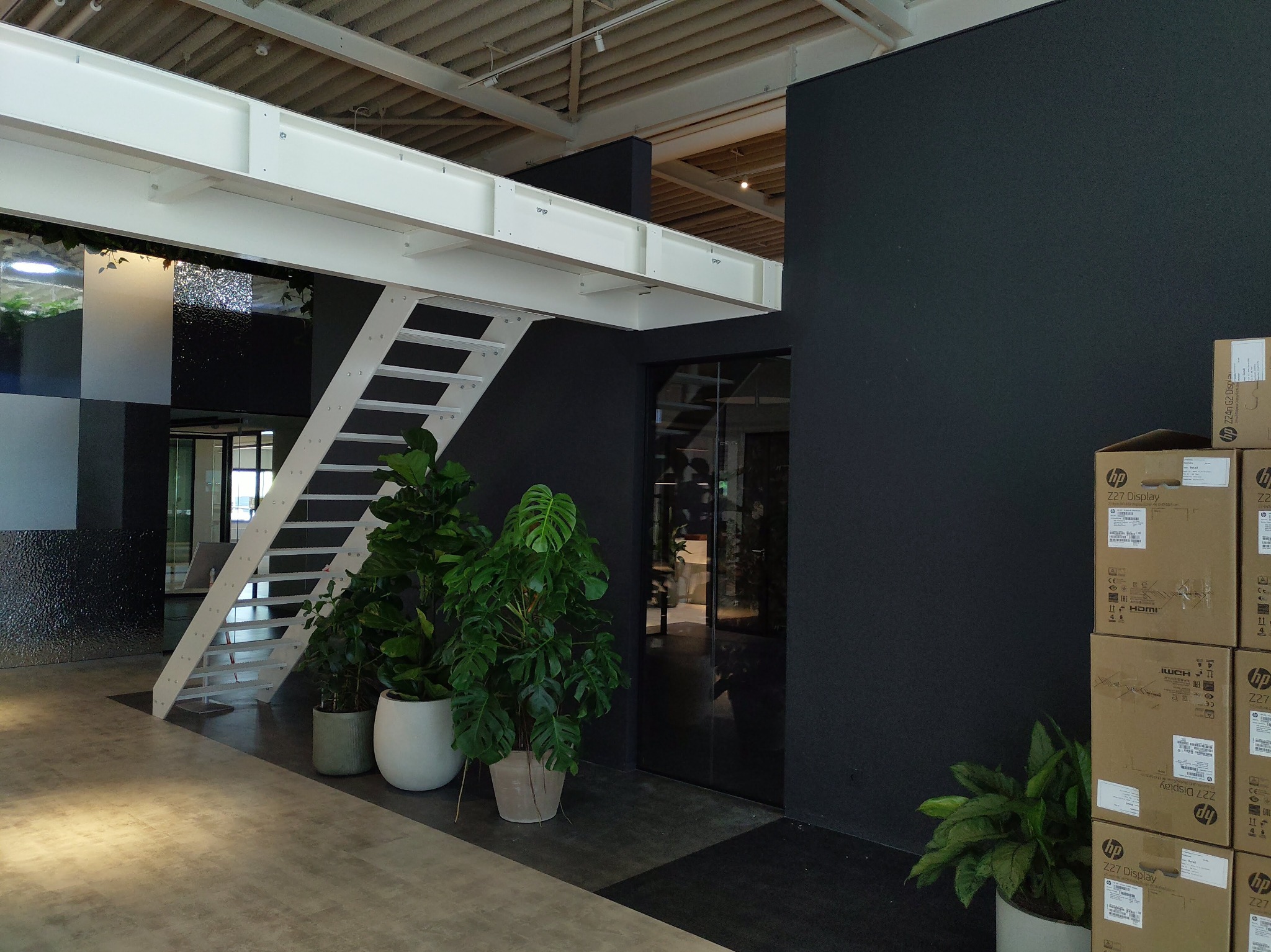 Nowoczesne wnętrze biurowe z czarną ścianą, białymi schodami na antresolę, roślinami doniczkowymi i stertą kartonów HP Z27 Display.