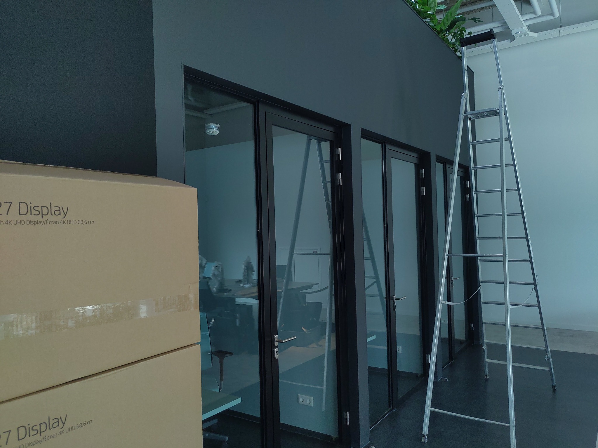 Wnętrze biura w trakcie przygotowań, z kartonami monitorów i rozstawioną aluminiową drabiną przy przeszklonych ściankach działowych.