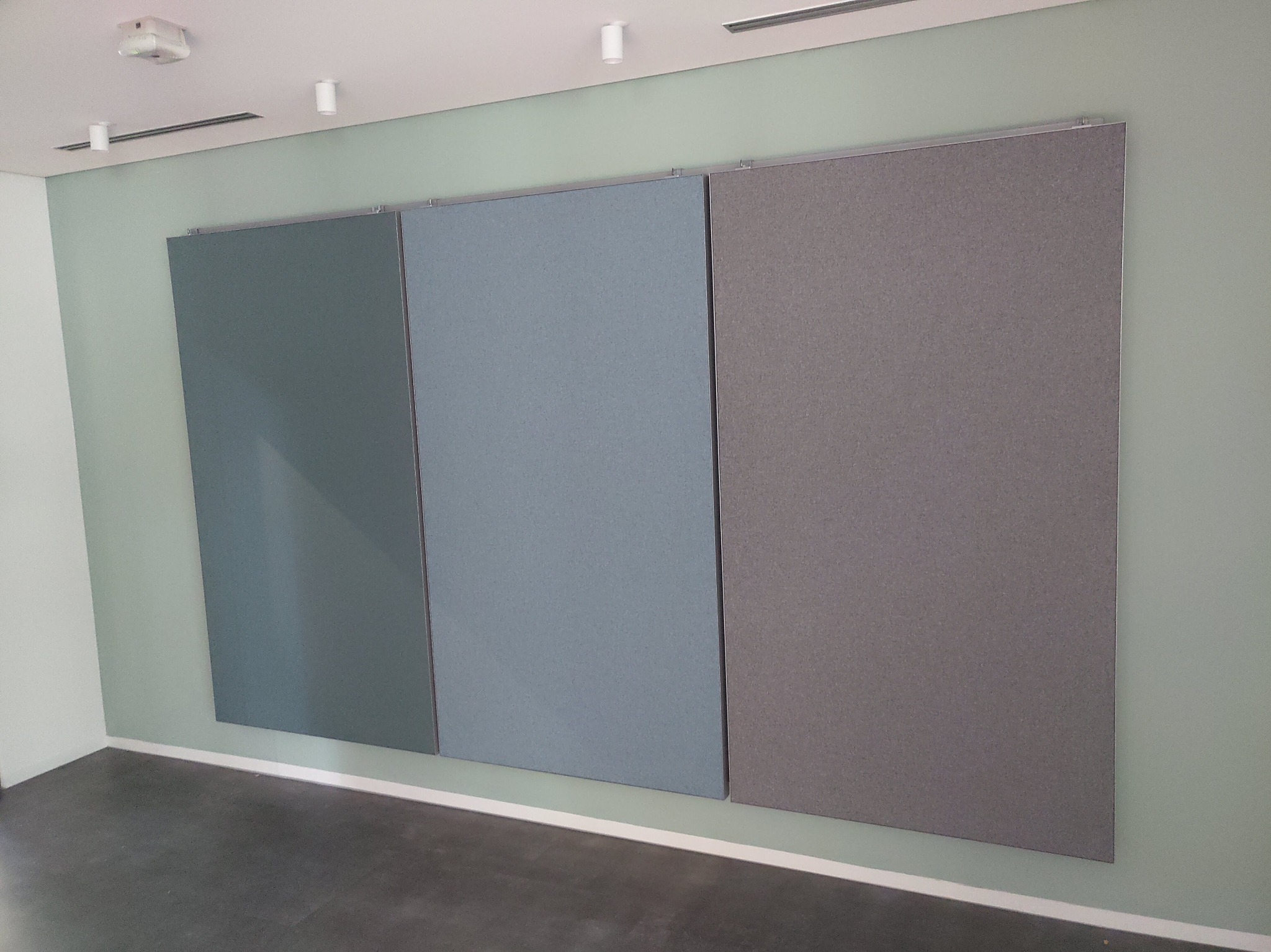 Trzy panele akustyczne w odcieniach szarości i błękitu zamontowane na ścianie w biurze, widoczny sufit z oświetleniem punktowym i wentylacją.