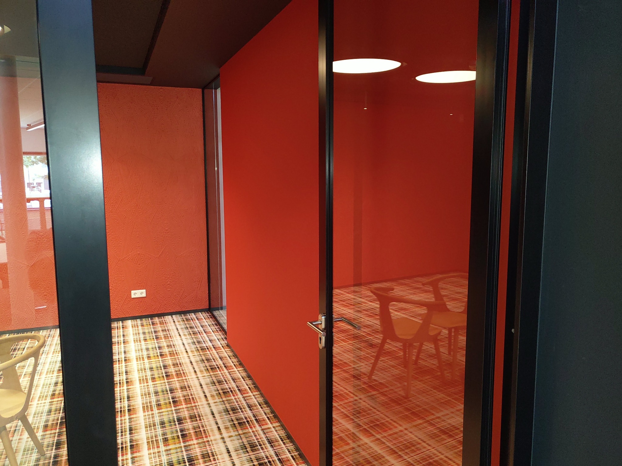 Pomarańczowy pokój biurowy z przeszklonymi ścianami i wzorzystą wykładziną, widok przez otwarte drzwi z czarną futryną.