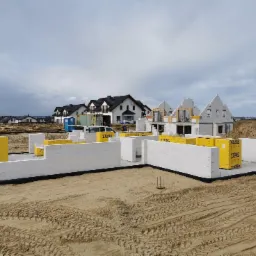 Wznoszone ściany fundamentowe z białych bloczków Ytong na piaszczystym gruncie, w tle budowa kolejnych domów jednorodzinnych.