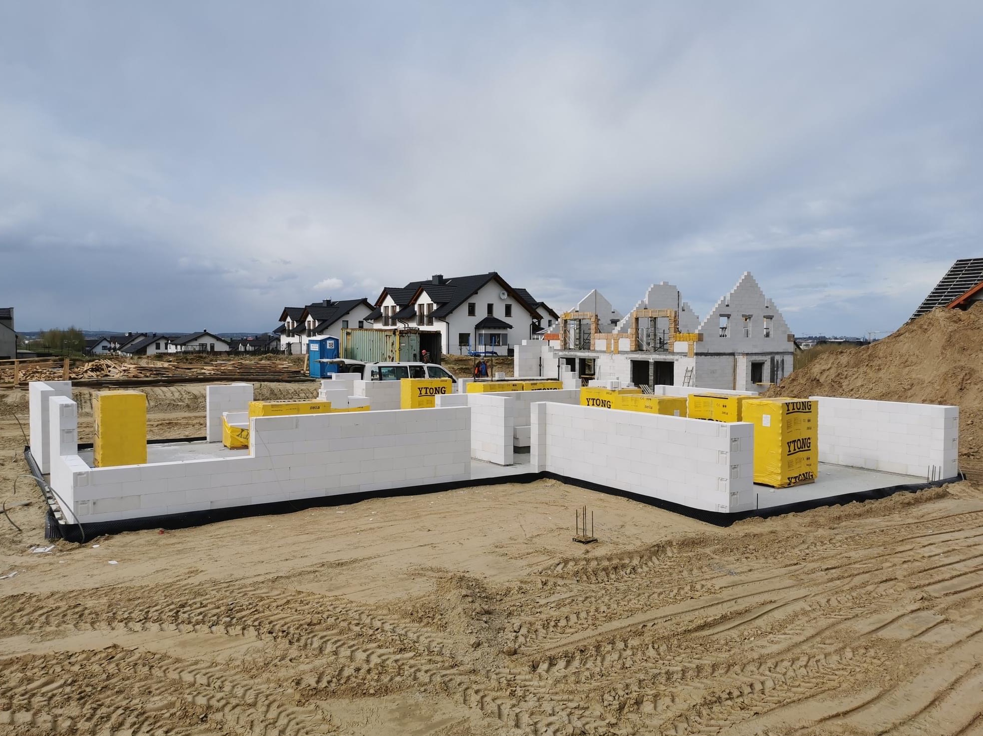Wznoszone ściany fundamentowe z białych bloczków Ytong na piaszczystym gruncie, w tle budowa kolejnych domów jednorodzinnych.