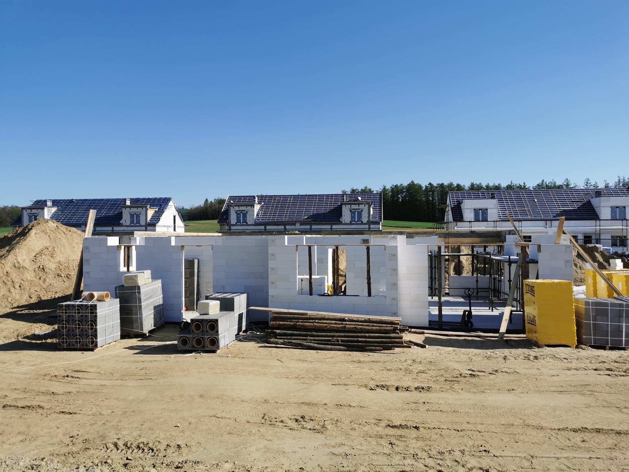 Budowa domów jednorodzinnych z białych bloczków, widoczne ściany w stanie surowym otwartym, ułożone bloczki i drewno budowlane, w tle domy z zamontowaną dachówką i panele fotowoltaiczne.