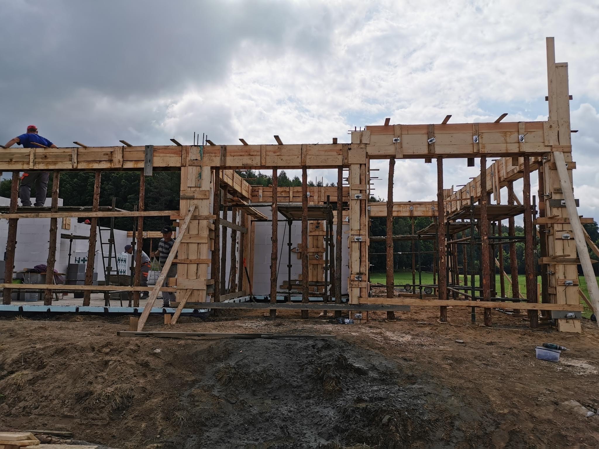 Wznoszenie szalunku drewnianego pod budowę domu jednorodzinnego, widoczne zbrojenie, bloczki fundamentowe i dwóch pracowników w tle, pochmurne niebo.