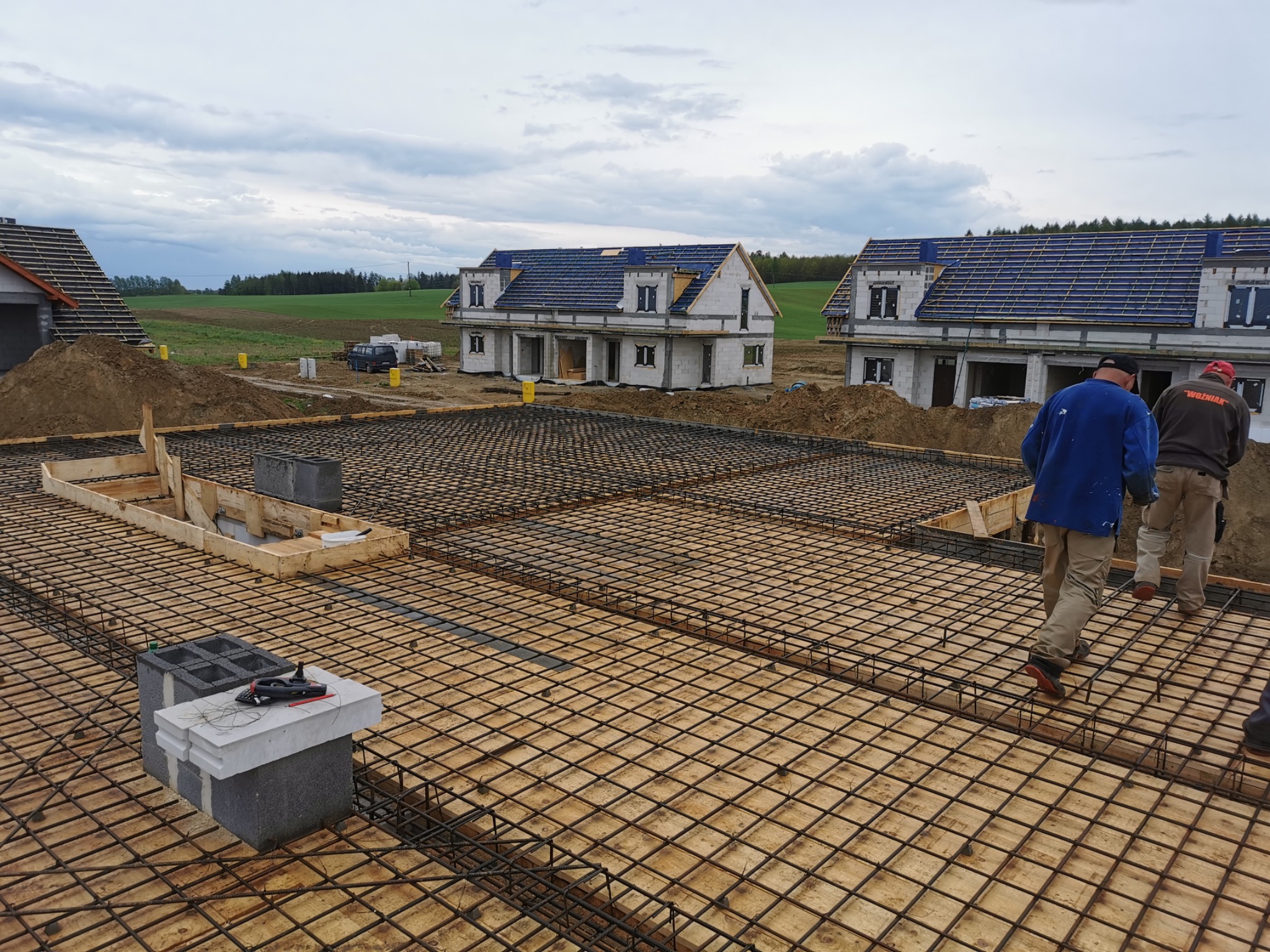Zbrojenie płyty fundamentowej na drewnianym szalunku, w tle wznoszące się domy jednorodzinne z niebieską dachówką i widok na pole uprawne pod pochmurnym niebem.
