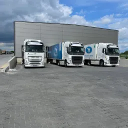 Trzy białe ciężarówki Volvo z logo firmy transportowej Chenczke Logistic zaparkowane przed szarą halą magazynową w pochmurny dzień.