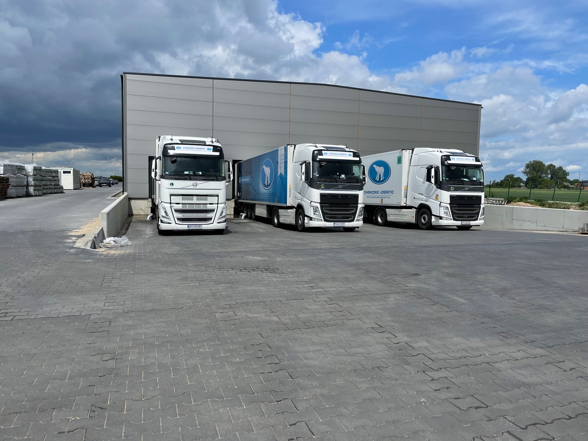 Trzy białe ciężarówki Volvo z logo firmy transportowej Chenczke Logistic zaparkowane przed szarą halą magazynową w pochmurny dzień.