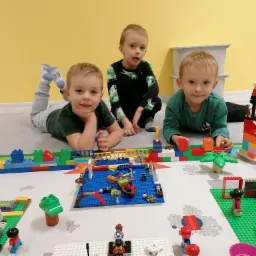 Trójka uśmiechniętych chłopców bawi się klockami Lego, tworząc miasteczko z portem i plażą na białej macie, widok z góry, żółta ściana w tle.