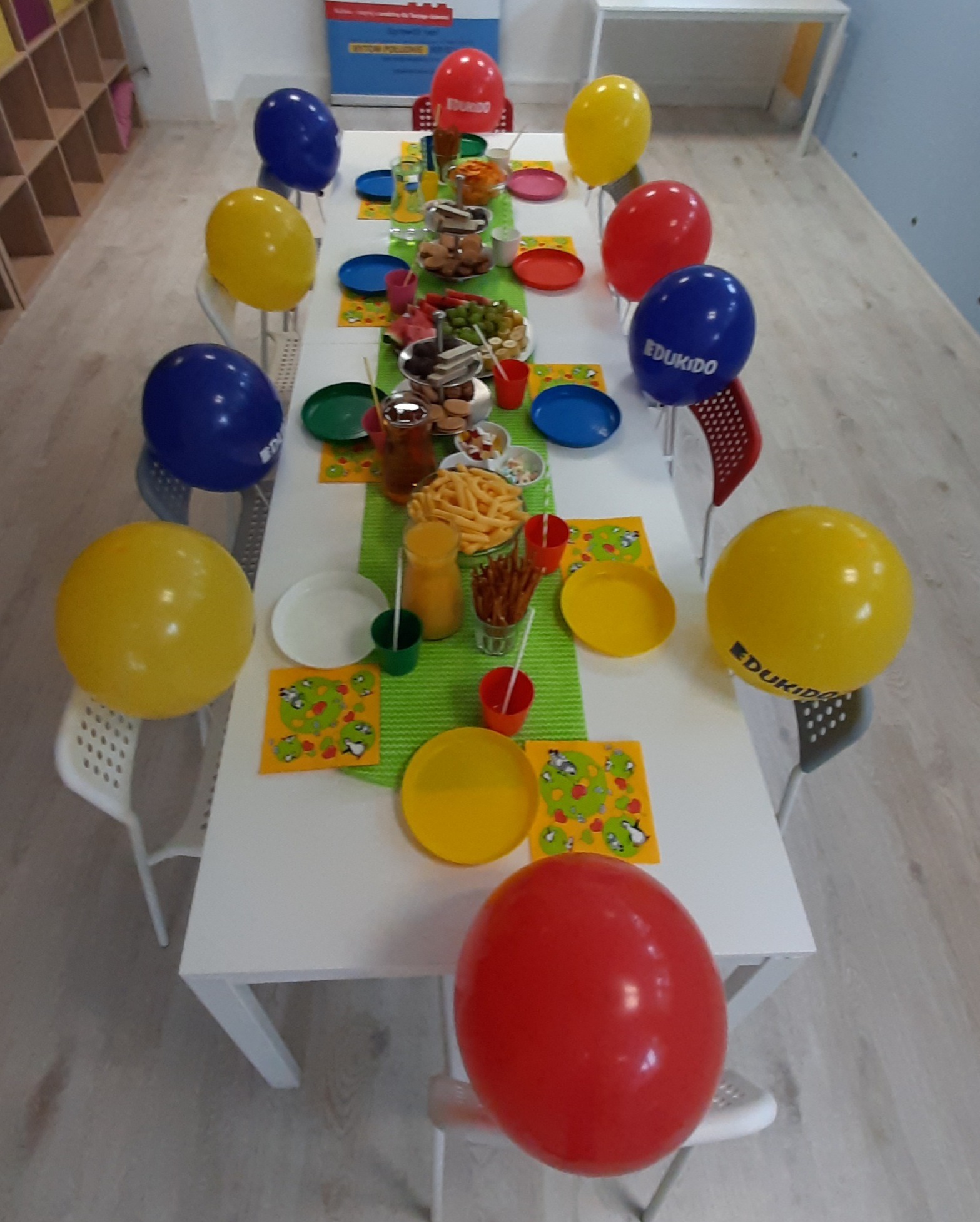 Stół udekorowany kolorowymi balonami z logo firmy, talerzami i przekąskami, przygotowany na przyjęcie dla dzieci.