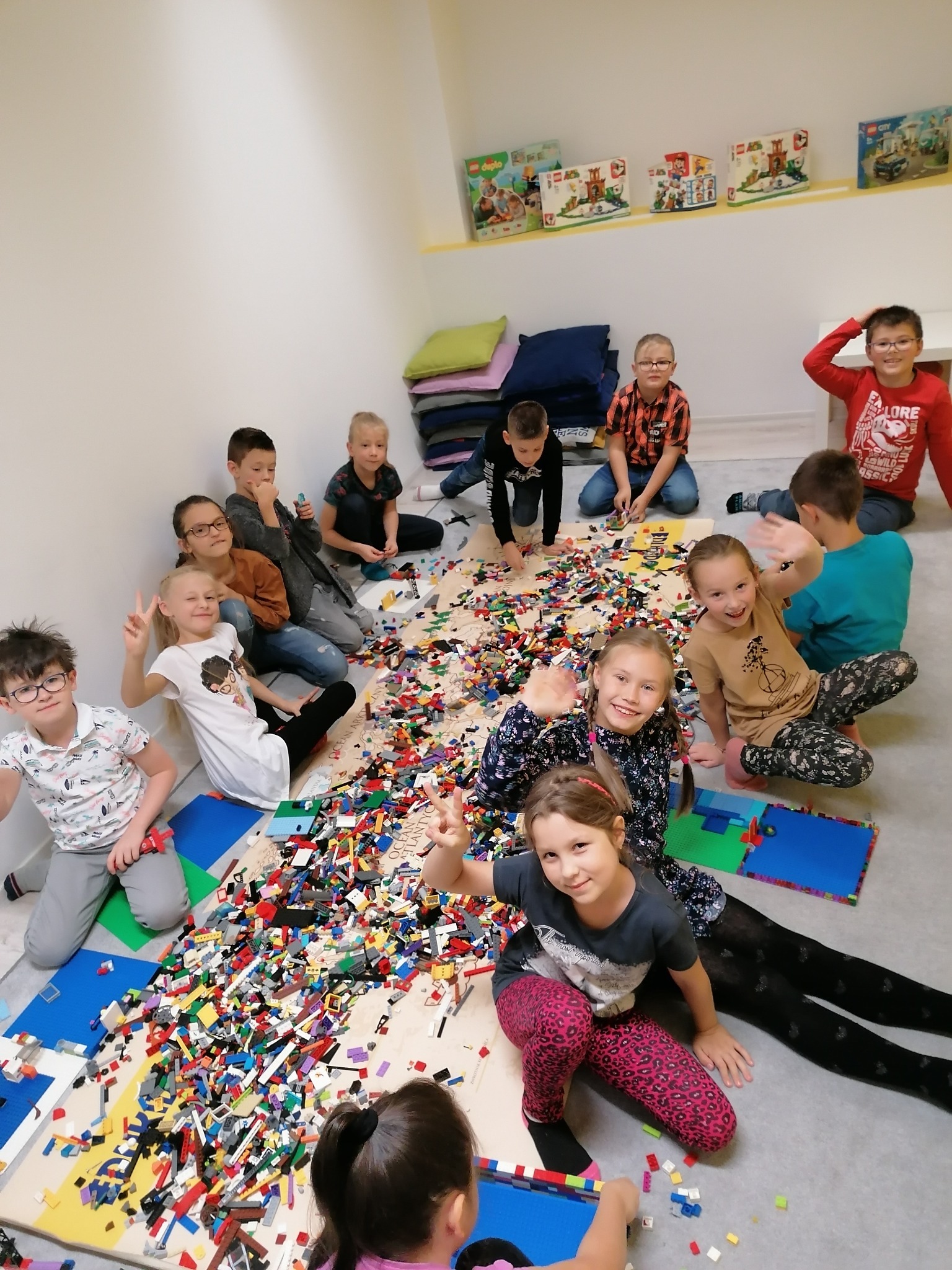 Grupa dzieci siedzi na podłodze otoczona kolorowymi klockami Lego, część z nich macha do kamery, a w tle widać półkę z pudełkami Lego.