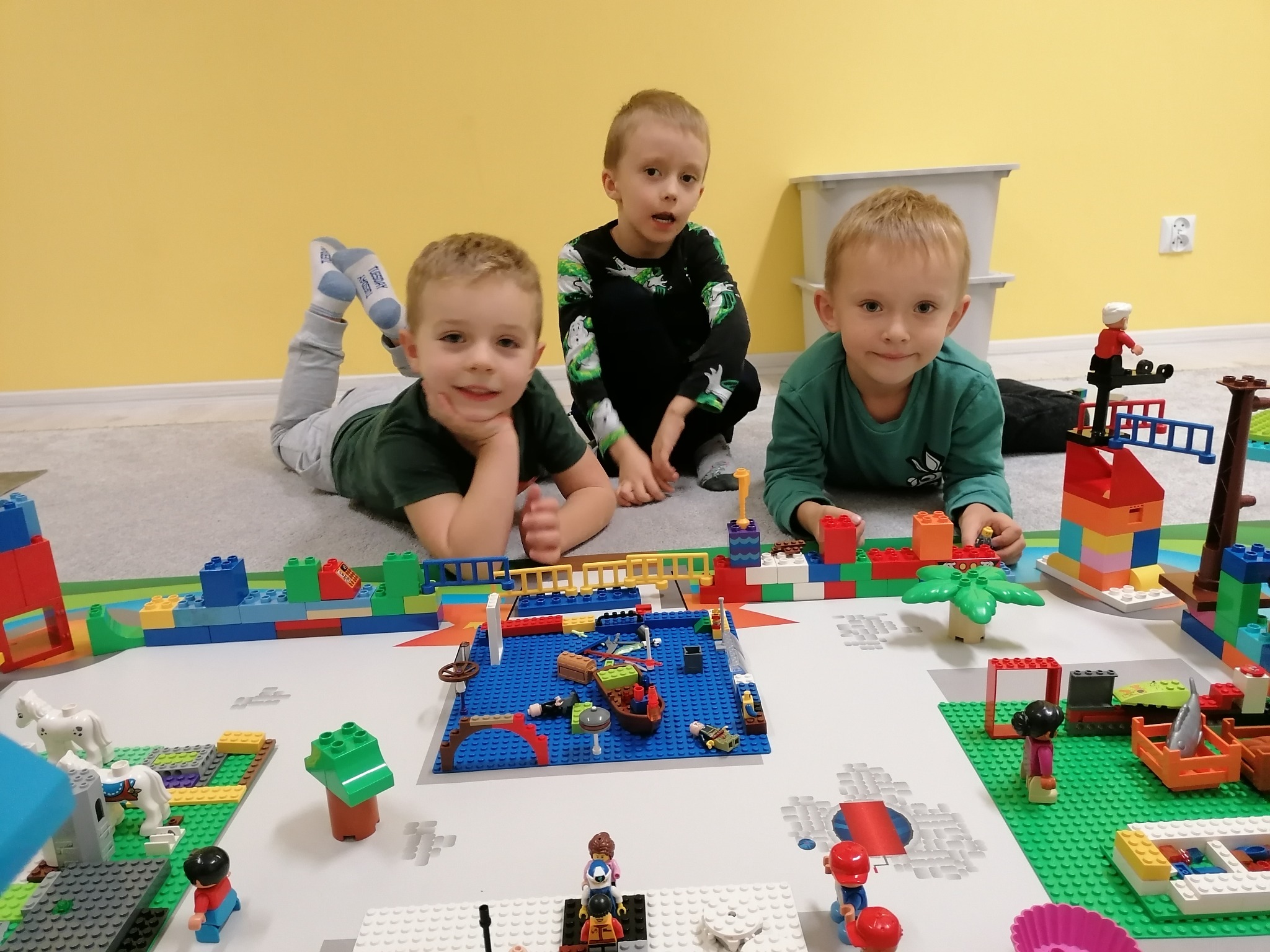 Trójka uśmiechniętych chłopców bawi się klockami Lego, tworząc miasteczko z portem i plażą na białej macie, widok z góry, żółta ściana w tle.