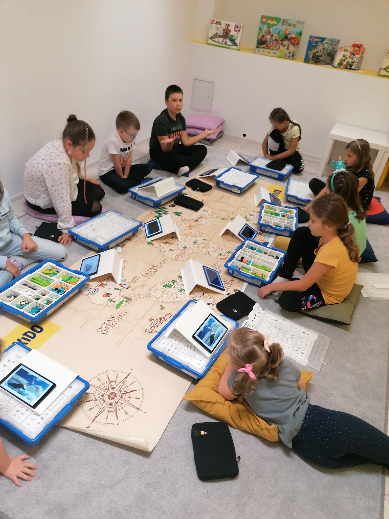 Grupa dzieci siedzi na podłodze wokół dużej mapy świata, korzystając z tabletów i zestawów edukacyjnych z klockami, w jasnym pomieszczeniu.