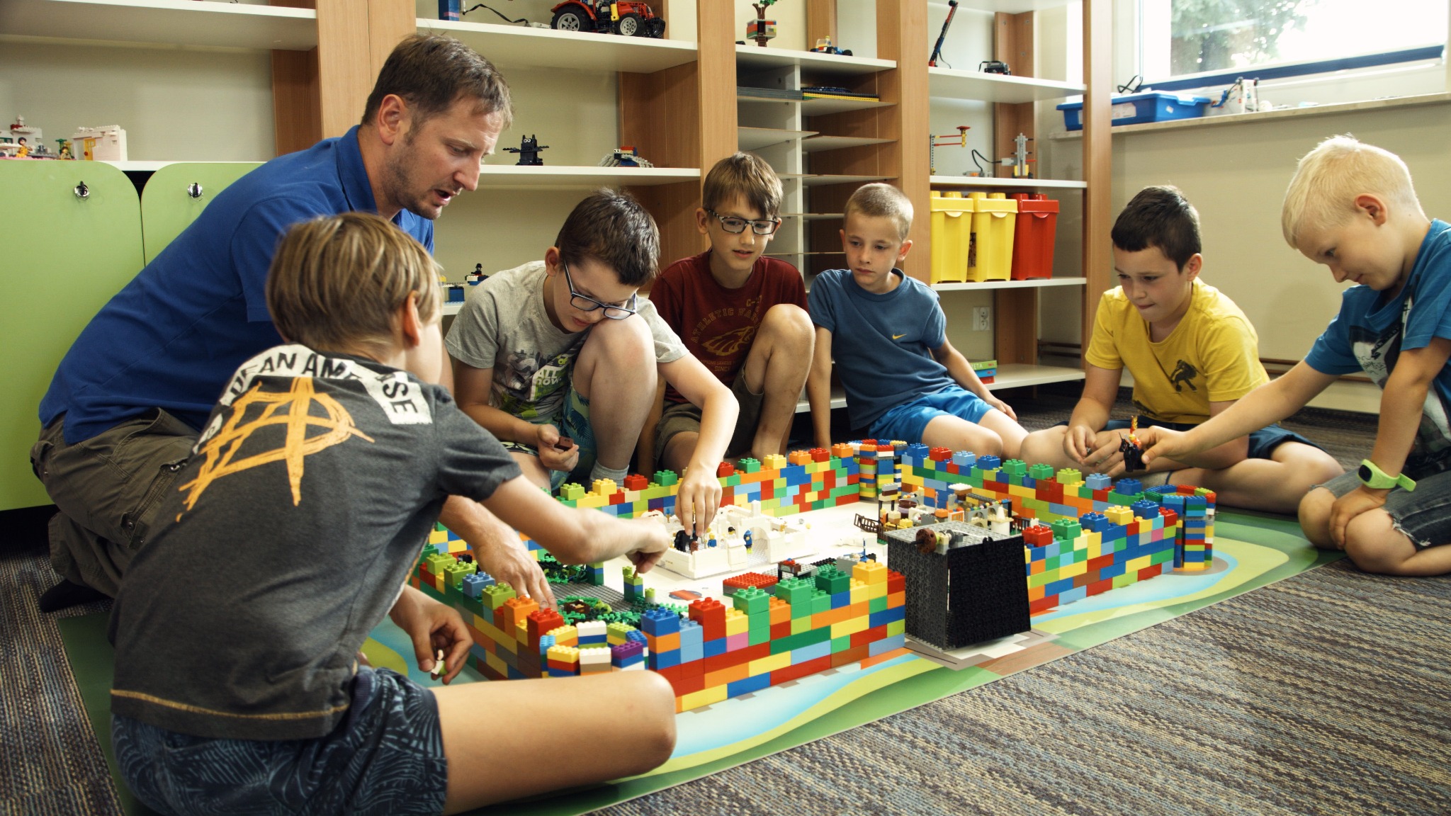 Grupa dzieci z opiekunem buduje miasto z kolorowych klocków na zielonej macie w jasnym pomieszczeniu z regałami pełnymi zabawek w tle.