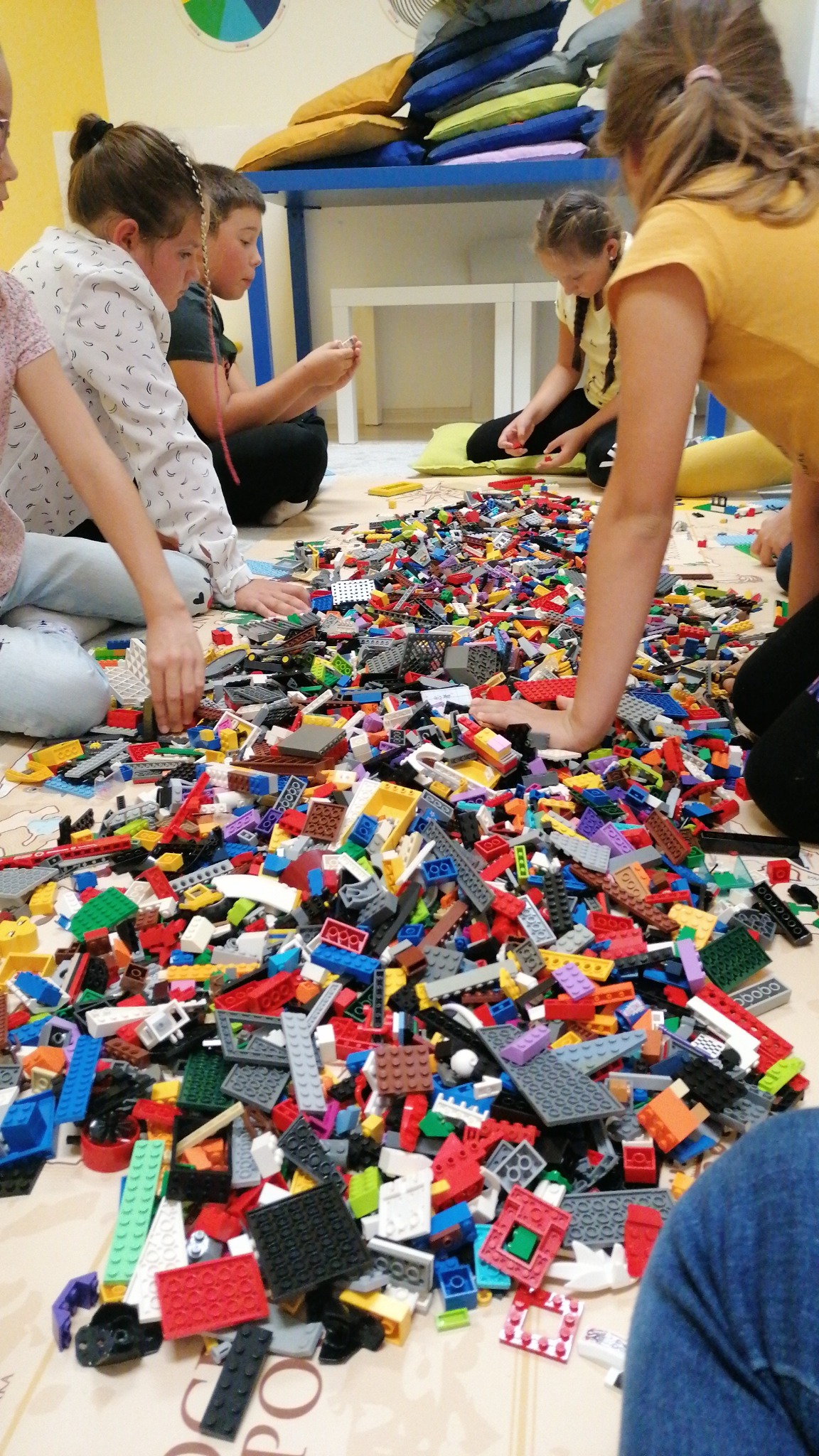 Grupa dzieci siedzi na podłodze wokół ogromnej sterty kolorowych klocków Lego, budując konstrukcje podczas zajęć grupowych.