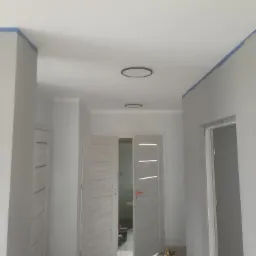 Korytarz w trakcie malowania: ściany w szarym kolorze, zabezpieczone niebieską taśmą malarską przy suficie, widoczne dwie nowoczesne lampy sufitowe i otwarte, białe drzwi z poziomymi przeszkleniami.