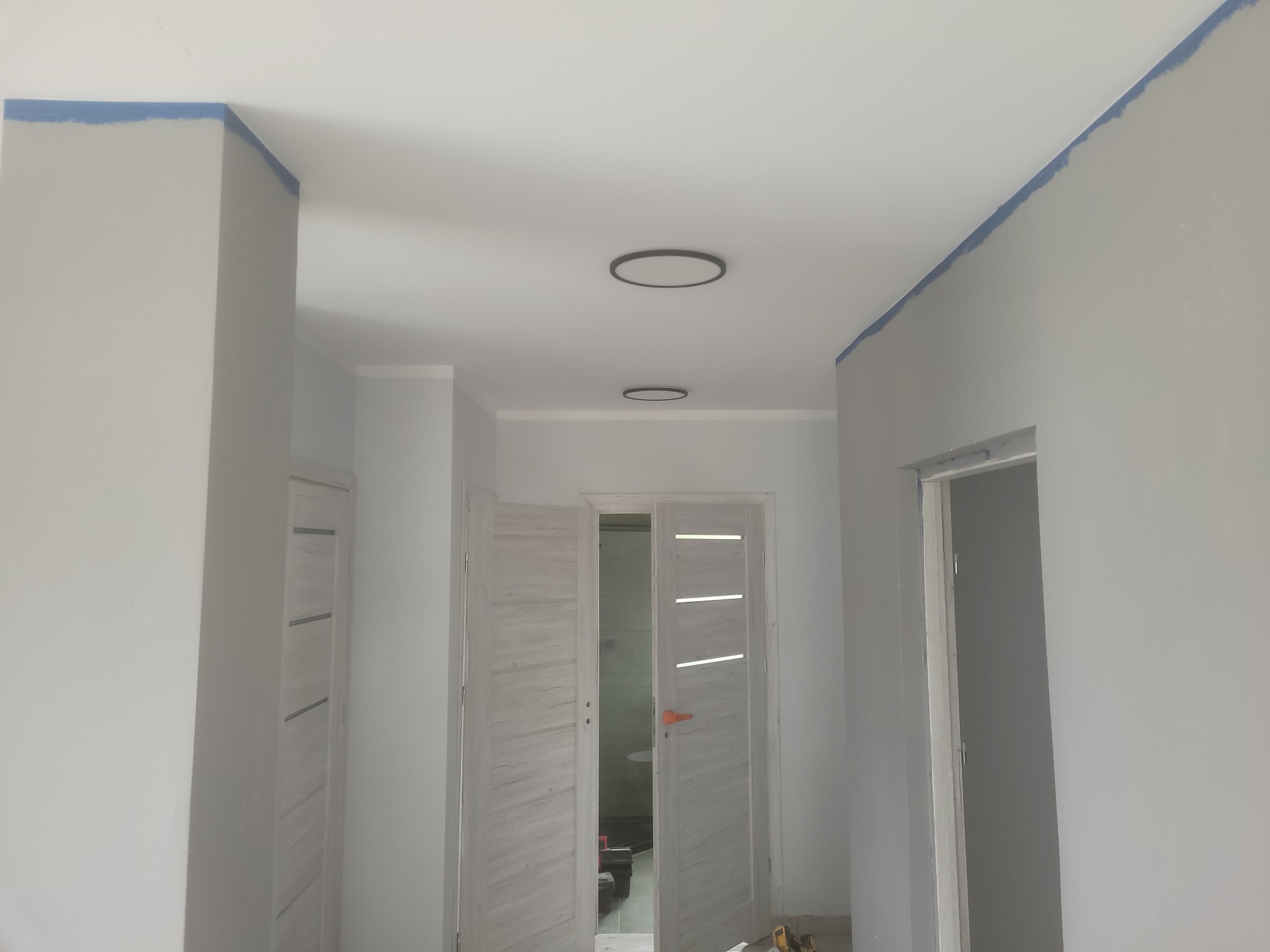 Korytarz w trakcie malowania: ściany w szarym kolorze, zabezpieczone niebieską taśmą malarską przy suficie, widoczne dwie nowoczesne lampy sufitowe i otwarte, białe drzwi z poziomymi przeszkleniami.