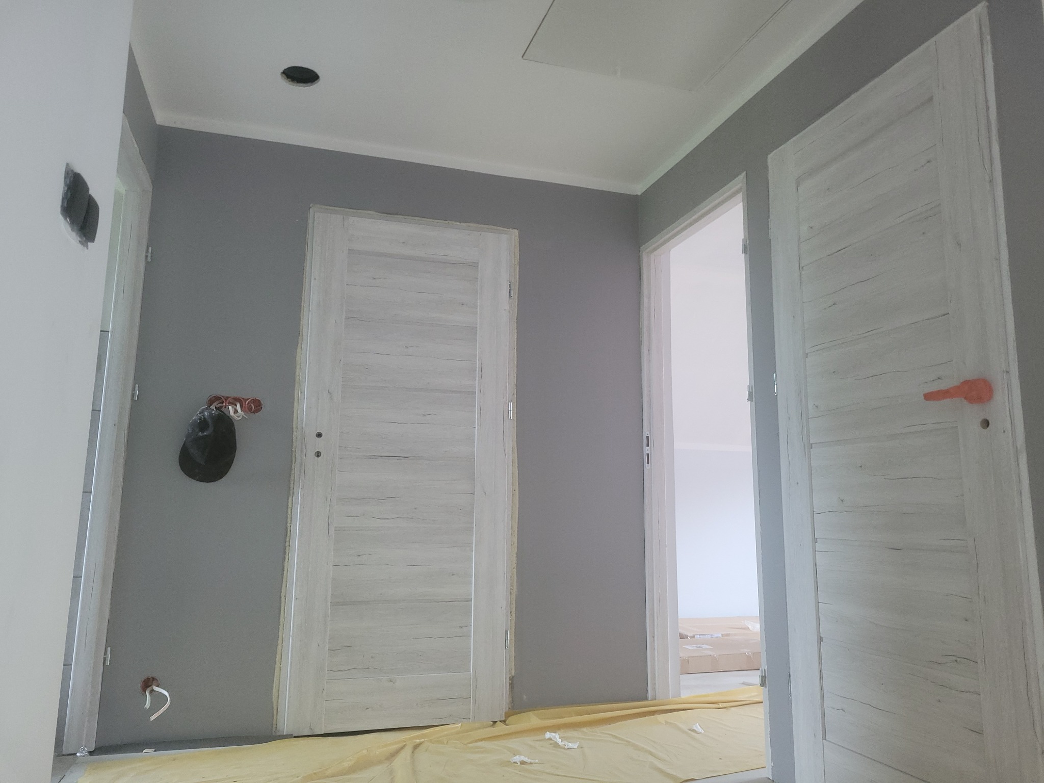 Wnętrze w trakcie prac wykończeniowych: szara ściana z dwiema zamontowanymi białymi drzwiami o strukturze drewna, podłoga zabezpieczona folią malarską, widoczne niedokończone instalacje elektryczne.