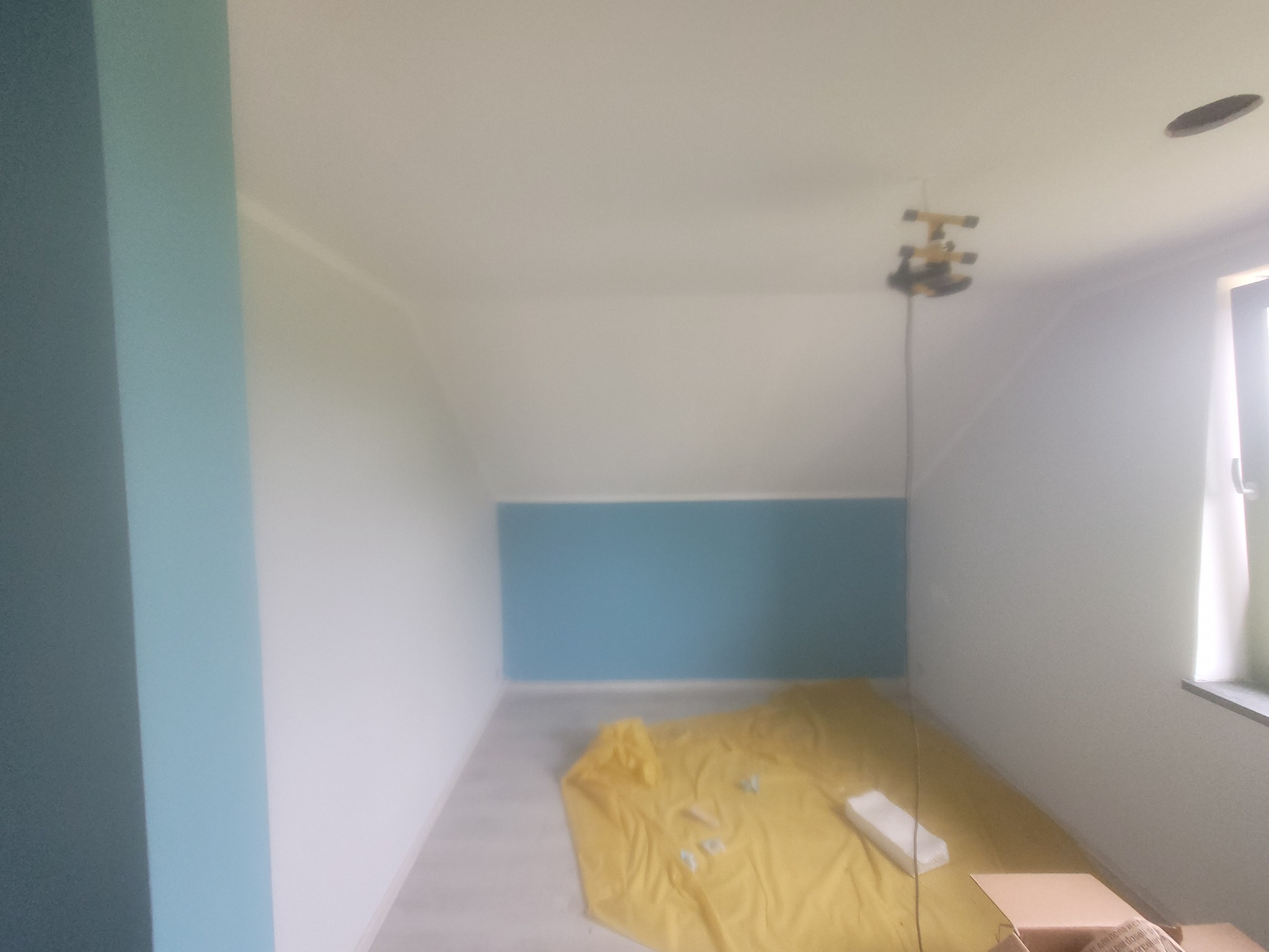 Poddasze po częściowym pomalowaniu: ściany białe i turkusowe, żółta folia zabezpieczająca podłogę, poziomica laserowa na suficie.