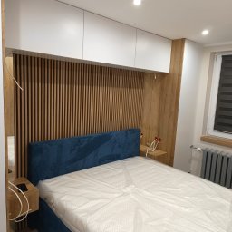 F. H. U. SONIA WIDAJEWICZ - Nowoczesna sypialnia z łóżkiem z niebieskim wezgłowiem, drewnianą ścianką z pionowych listew, białymi szafkami wiszącymi i drewnianymi szafkami nocnymi, częściowo zakrytym materacem folią ochronną.