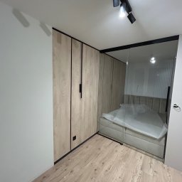 F. H. U. SONIA WIDAJEWICZ - Nowoczesna szafa wnękowa z drewnopodobnymi frontami i czarnymi detalami, przylegająca do szklanej ściany oddzielającej sypialnię z łóżkiem przykrytym folią ochronną.