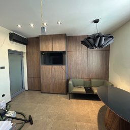 F. H. U. SONIA WIDAJEWICZ - Nowoczesne wnętrze z zabudową meblową z drewnopodobnej płyty, telewizorem wkomponowanym w szafę, szarą sofą, designerską lampą i okrągłym stołem. Na podłodze leży odkurzacz przemysłowy, a z sufitu...