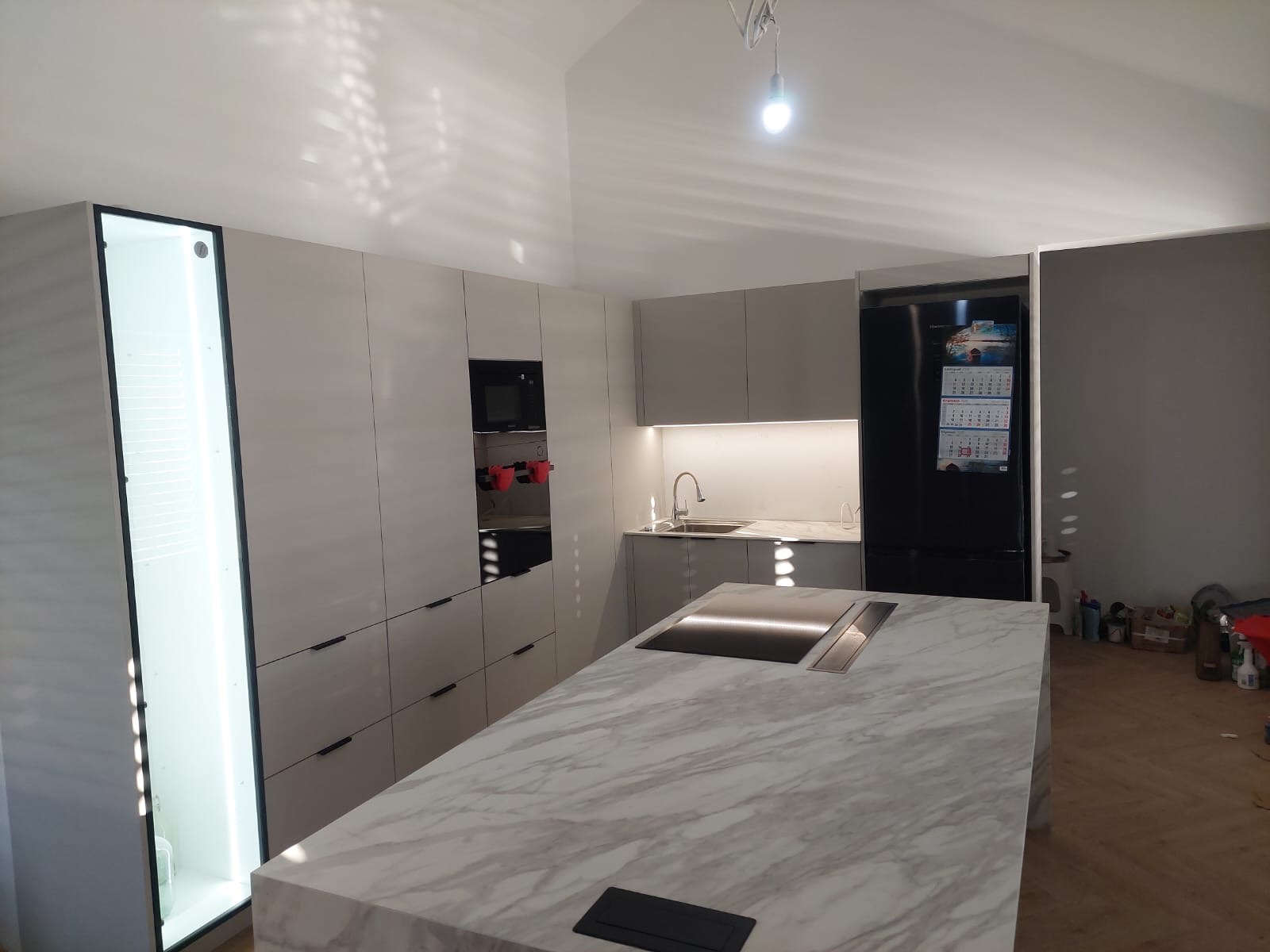 Nowoczesna kuchnia z wyspą z marmurowym blatem, zabudową w odcieniach szarości i oświetleniem LED, widoczna czarna lodówka z kalendarzem.