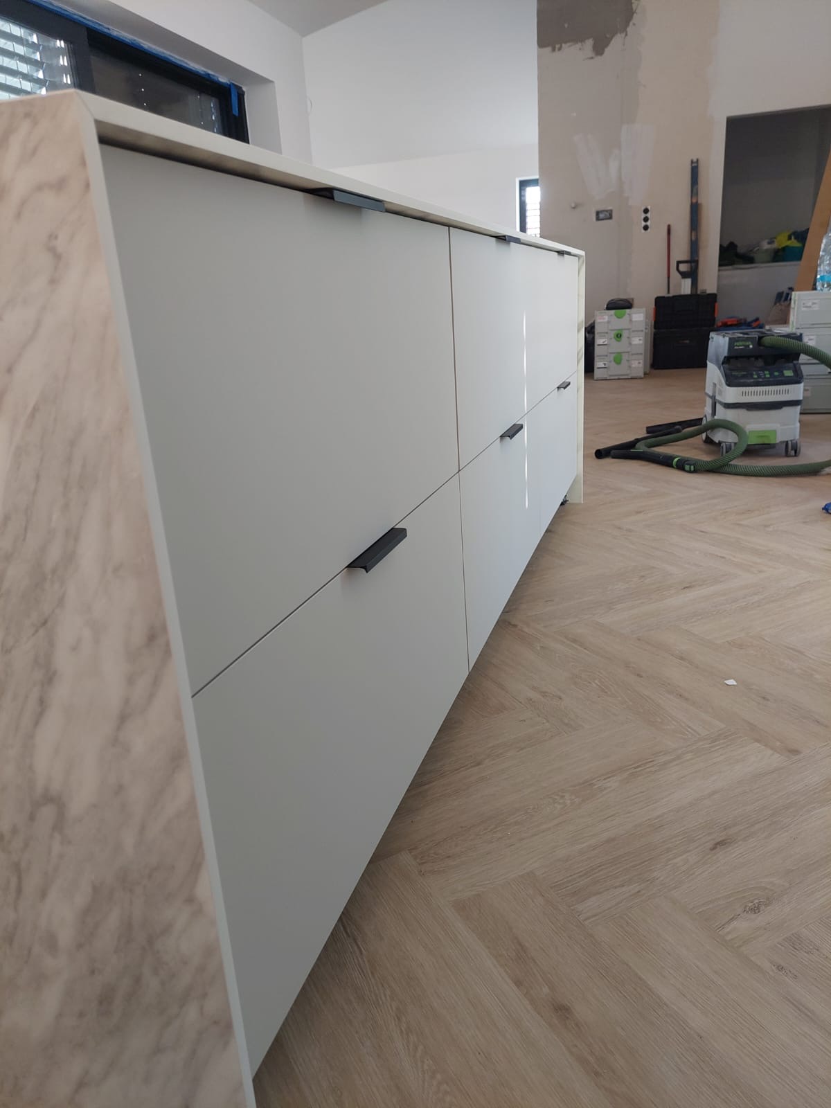 Fragment zabudowy kuchennej z szarymi frontami i blatem imitującym marmur, widoczny od strony bocznej, z odkurzaczem przemysłowym w tle i parkietem ułożonym w jodełkę.